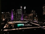 A nighttime cityscape featuring a large, prominent building with illuminated purple and white lights. Below, a large sign reading 'TORONTO' is lit up in green and white. The area is surrounded by other buildings with numerous lit windows and city lights, contributing to an urban evening atmosphere.