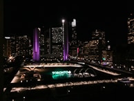 A nighttime cityscape featuring a large, prominent building with illuminated purple and white lights. Below, a large sign reading 'TORONTO' is lit up in green and white. The area is surrounded by other buildings with numerous lit windows and city lights, contributing to an urban evening atmosphere.