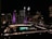 A nighttime cityscape featuring a large, prominent building with illuminated purple and white lights. Below, a large sign reading 'TORONTO' is lit up in green and white. The area is surrounded by other buildings with numerous lit windows and city lights, contributing to an urban evening atmosphere.