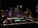 A nighttime cityscape featuring a large, prominent building with illuminated purple and white lights. Below, a large sign reading 'TORONTO' is lit up in green and white. The area is surrounded by other buildings with numerous lit windows and city lights, contributing to an urban evening atmosphere.