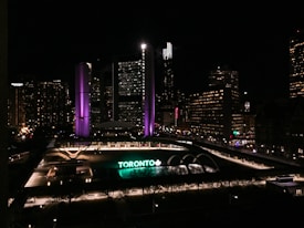 A nighttime cityscape featuring a large, prominent building with illuminated purple and white lights. Below, a large sign reading 'TORONTO' is lit up in green and white. The area is surrounded by other buildings with numerous lit windows and city lights, contributing to an urban evening atmosphere.
