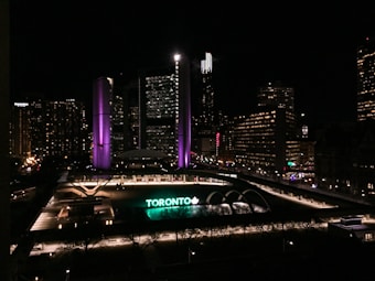 A nighttime cityscape featuring a large, prominent building with illuminated purple and white lights. Below, a large sign reading 'TORONTO' is lit up in green and white. The area is surrounded by other buildings with numerous lit windows and city lights, contributing to an urban evening atmosphere.