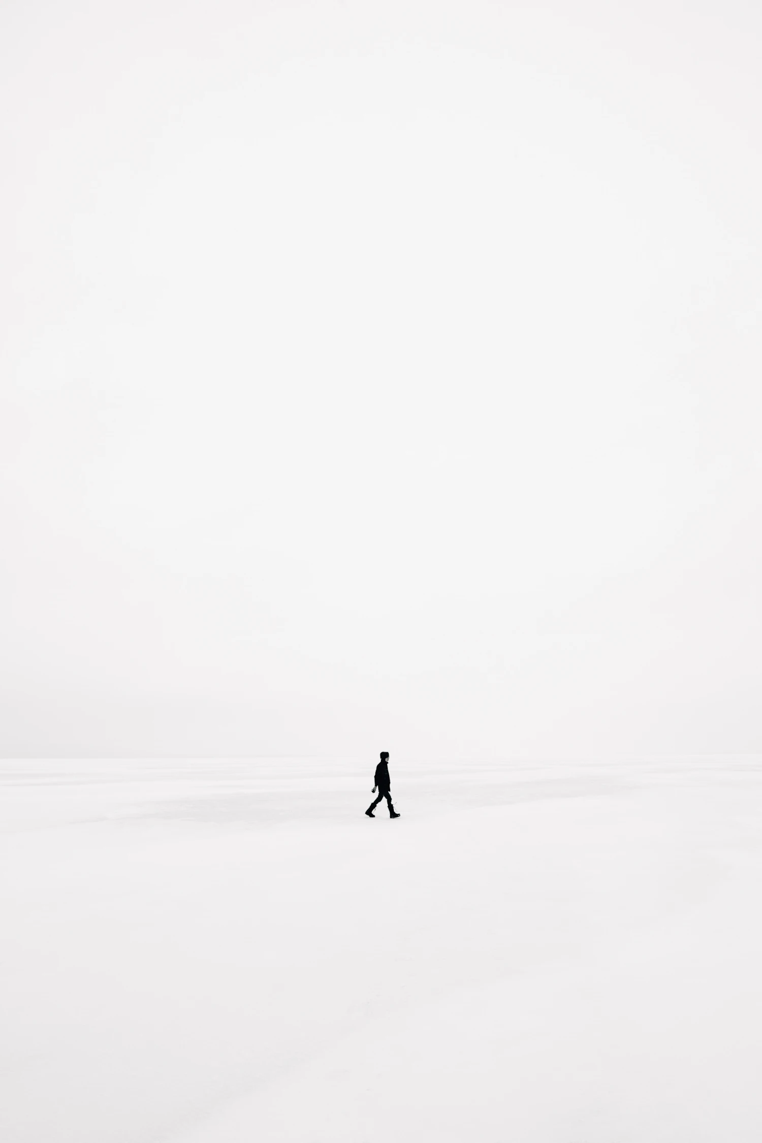 A single small figure walking across an empty snowfield under a white sky