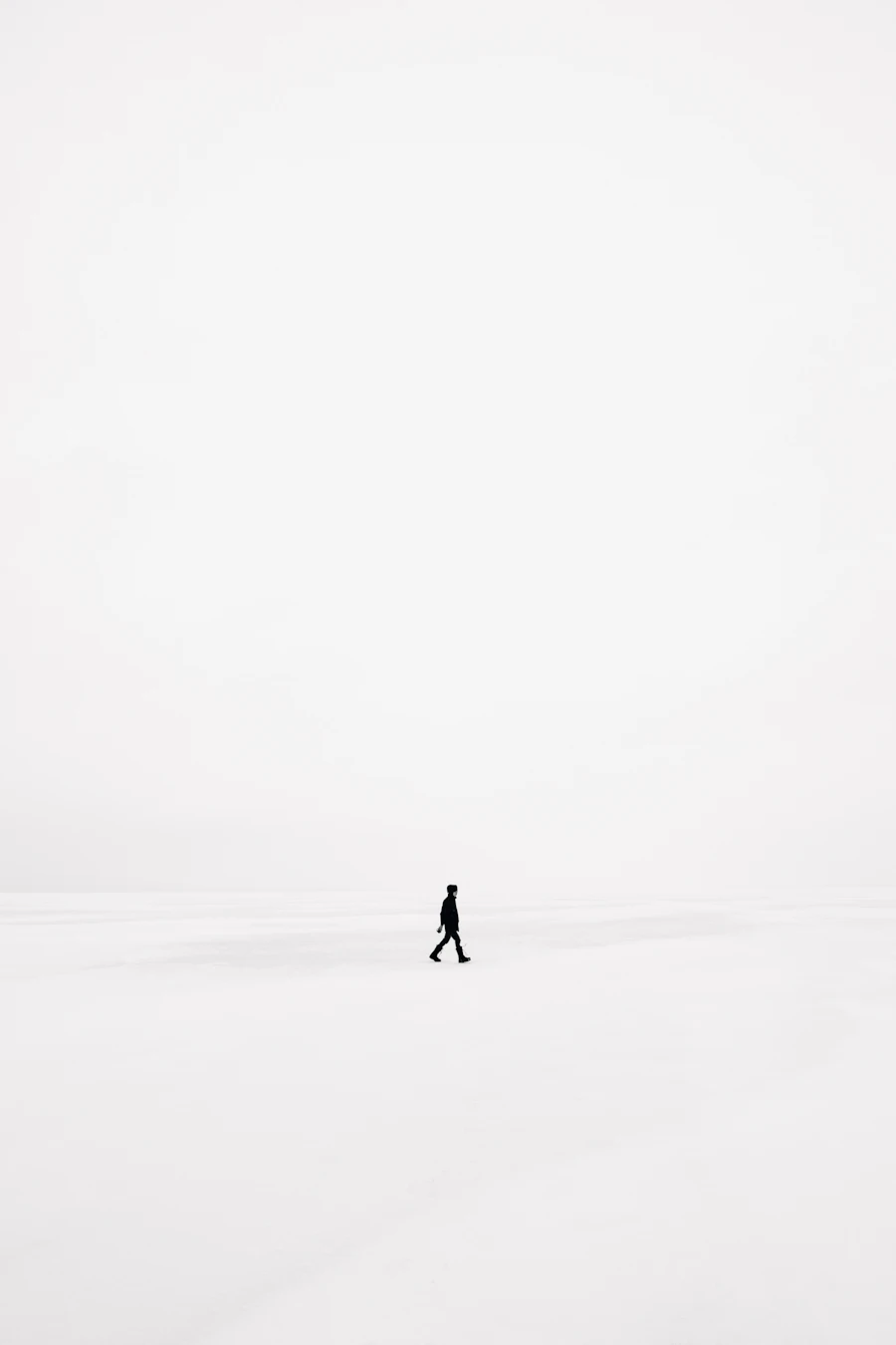 A single small figure walking across an empty snowfield under a white sky