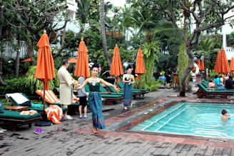 A lush outdoor setting with a swimming pool surrounded by green foliage and orange umbrellas. People are relaxing on loungers, with some individuals standing and interacting. A performer in traditional attire is also visible.