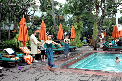A lush outdoor setting with a swimming pool surrounded by green foliage and orange umbrellas. People are relaxing on loungers, with some individuals standing and interacting. A performer in traditional attire is also visible.