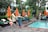 A lush outdoor setting with a swimming pool surrounded by green foliage and orange umbrellas. People are relaxing on loungers, with some individuals standing and interacting. A performer in traditional attire is also visible.