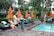 A lush outdoor setting with a swimming pool surrounded by green foliage and orange umbrellas. People are relaxing on loungers, with some individuals standing and interacting. A performer in traditional attire is also visible.