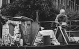 An older man is engaged in some work on a boat, surrounded by various items including a bicycle, rope, and wooden structures. The scene has an industrial feel with an abundance of metallic and wooden textures.