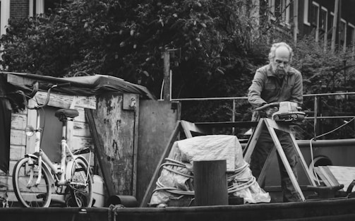 An older man is engaged in some work on a boat, surrounded by various items including a bicycle, rope, and wooden structures. The scene has an industrial feel with an abundance of metallic and wooden textures.