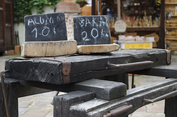 A rustic scene with two old wooden beams displaying chalkboard signs. The signs have hand-written text: 'ALUM 101' on one and 'BALA 20%' on the other. These are positioned outdoors, with a market or shop stall in the blurred background holding various products.