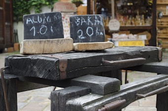 A rustic scene with two old wooden beams displaying chalkboard signs. The signs have hand-written text: 'ALUM 101' on one and 'BALA 20%' on the other. These are positioned outdoors, with a market or shop stall in the blurred background holding various products.