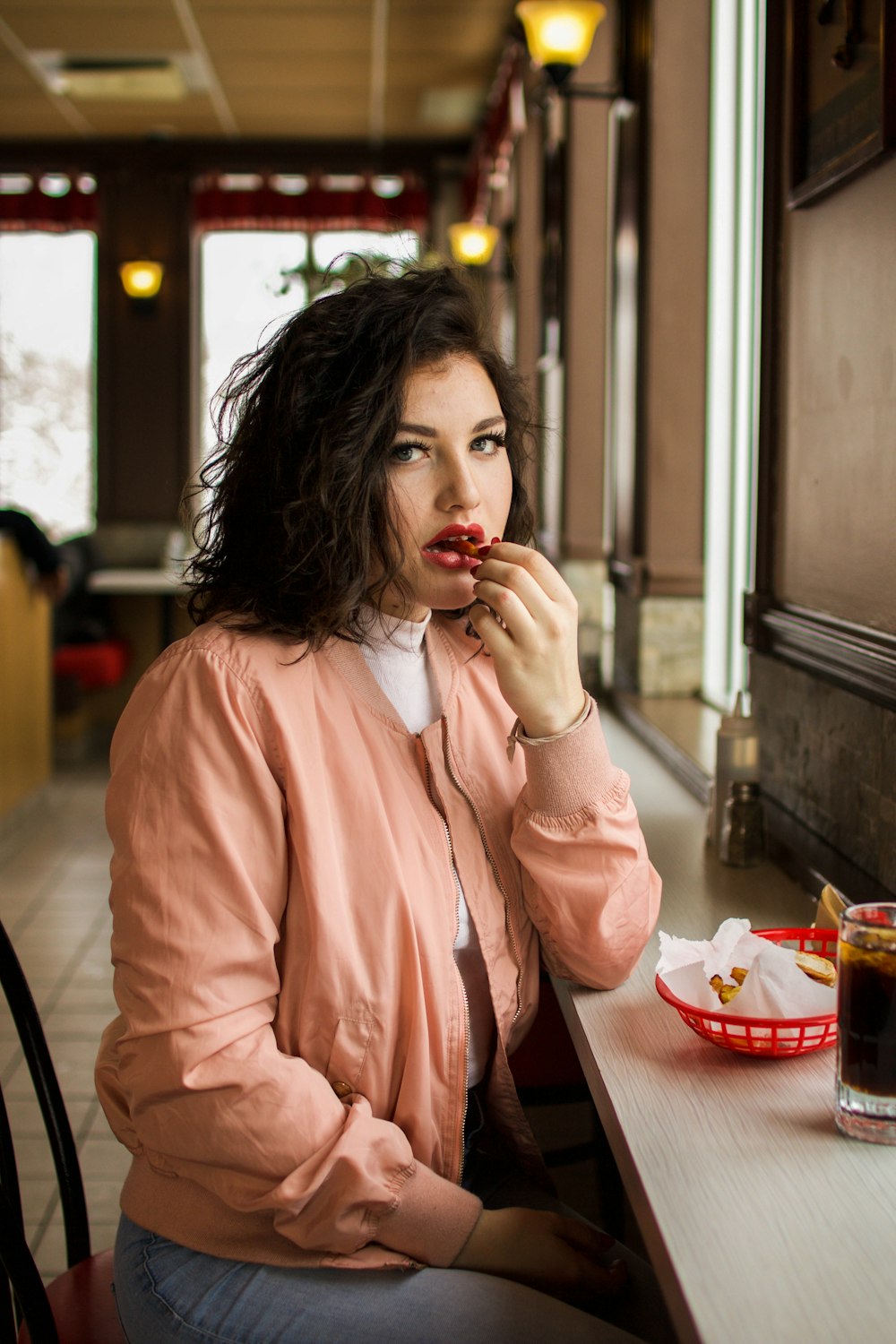 A woman in a pink jacket sits at a counter, eating and looking at the camera