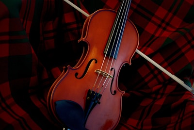 A violin resting on a red and black plaid fabric, with a bow placed across the instrument. The warm brown tones of the wooden violin contrast with the vibrant pattern of the cloth.
