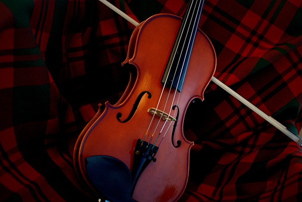 A violin resting on a red and black plaid fabric, with a bow placed across the instrument. The warm brown tones of the wooden violin contrast with the vibrant pattern of the cloth.