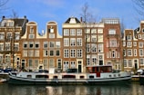 A canal boat is docked in front of a row of traditional Dutch-style buildings with gabled roofs. The buildings are tall and narrow, featuring large windows and brick facades. Leafless trees are present, indicating a winter or early spring setting. Cars are parked along the street beside the canal.