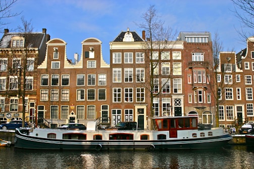 A canal boat is docked in front of a row of traditional Dutch-style buildings with gabled roofs. The buildings are tall and narrow, featuring large windows and brick facades. Leafless trees are present, indicating a winter or early spring setting. Cars are parked along the street beside the canal.