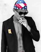 A person wearing sunglasses and a bandana with an American flag pattern is smoking a cigarette. They have a partially buttoned black blazer over their bare chest, with a package of cigarettes or similar item in the pocket.