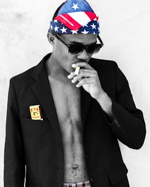 A person wearing sunglasses and a bandana with an American flag pattern is smoking a cigarette. They have a partially buttoned black blazer over their bare chest, with a package of cigarettes or similar item in the pocket.
