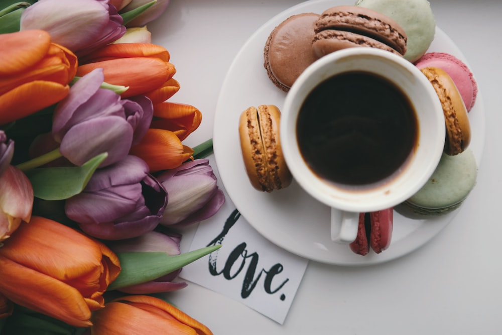 A black coffee in a small cup on top of a plate, surrounded by flowers and snacks.