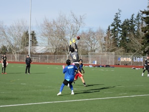 Several athletes on a sports field are playing football. One player is leaping into the air to catch a football, while others look on. The field is surrounded by trees and a wire fence. The weather seems clear and sunny.