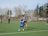Several athletes on a sports field are playing football. One player is leaping into the air to catch a football, while others look on. The field is surrounded by trees and a wire fence. The weather seems clear and sunny.