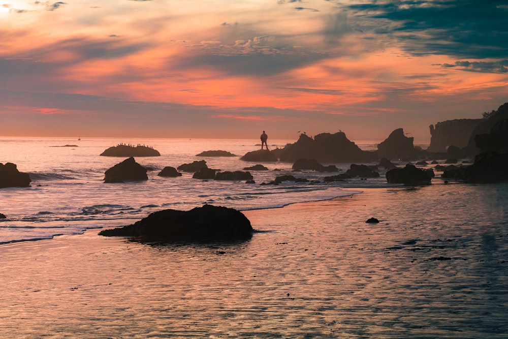 silhouette person standing on seashore rock during daytime