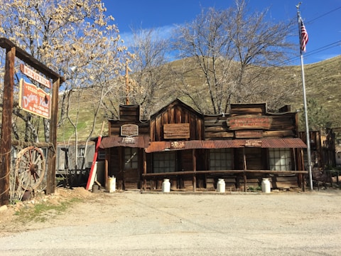 A rustic wooden building with an old-fashioned Western architectural style stands prominently in the scene. The structure features several signs with weathered, vintage text. Nearby, an American flag is displayed on a pole. The landscape includes bare-branched trees and hills in the background under a clear blue sky. A large sign with Western-style lettering is visible to the left, and a couple of old milk cans are placed in front of the building.