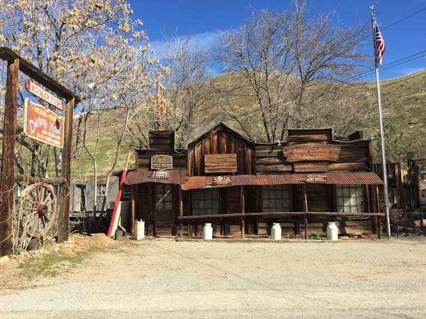 A rustic wooden building with an old-fashioned Western architectural style stands prominently in the scene. The structure features several signs with weathered, vintage text. Nearby, an American flag is displayed on a pole. The landscape includes bare-branched trees and hills in the background under a clear blue sky. A large sign with Western-style lettering is visible to the left, and a couple of old milk cans are placed in front of the building.
