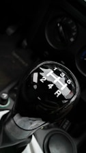 A close-up view of a manual car gear shift knob displaying the gear positions. The background includes parts of the car console, with visible circular controls and buttons.