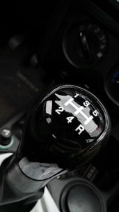 A close-up view of a manual car gear shift knob displaying the gear positions. The background includes parts of the car console, with visible circular controls and buttons.