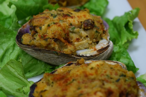 A close-up view of baked and stuffed clams resting on a bed of fresh green lettuce. The clams appear crispy and golden-brown with visible herbs and breadcrumbs on top, offering a savory and appetizing appearance.