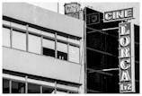 A black and white photograph of a building facade featuring large windows and a vertical sign. The sign displays the words 'CINE LORCA 1/2' with bold, retro-style lettering. The structure gives a slightly industrial look with a combination of concrete and glass elements.
