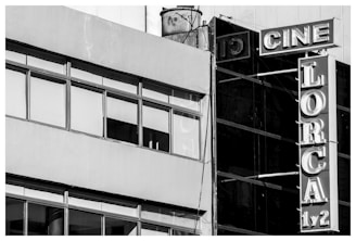 A black and white photograph of a building facade featuring large windows and a vertical sign. The sign displays the words 'CINE LORCA 1/2' with bold, retro-style lettering. The structure gives a slightly industrial look with a combination of concrete and glass elements.