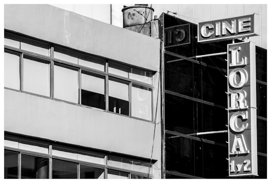 A black and white photograph of a building facade featuring large windows and a vertical sign. The sign displays the words 'CINE LORCA 1/2' with bold, retro-style lettering. The structure gives a slightly industrial look with a combination of concrete and glass elements.