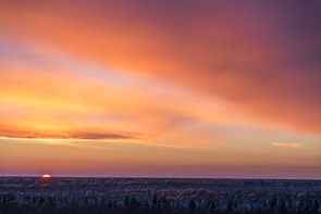 A vibrant sunset with a sky full of warm orange, pink, and purple hues spreads across the horizon. The sun is just visible above a dense forest line, creating a serene and picturesque scene.