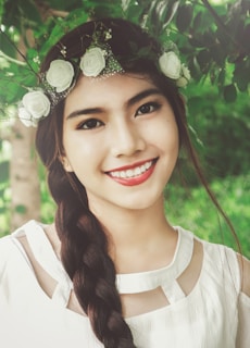A woman with a floral headpiece consisting of white roses and small green leaves poses against a backdrop of lush foliage. Her long braided hair drapes over her shoulder, and she wears a white dress with cutout details near the neckline. The expression on her face is warm and inviting, accentuated by her bright smile and dark eyes.