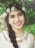 A woman with a floral headpiece consisting of white roses and small green leaves poses against a backdrop of lush foliage. Her long braided hair drapes over her shoulder, and she wears a white dress with cutout details near the neckline. The expression on her face is warm and inviting, accentuated by her bright smile and dark eyes.