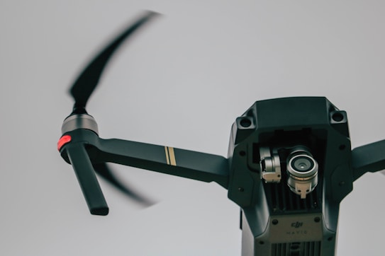 A close-up view of a drone with spinning propellers. The focus is on the camera and rotary parts of the drone, showcasing its mechanical design. The background is a neutral, overcast sky, emphasizing the flying device.