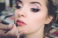 A close-up of a woman having makeup applied to her lips. Her eyes are accentuated with dark eyeliner and pink eye shadow. The focus is on her face, especially the eyes and lips, while the background is blurred, suggesting a makeup studio environment.