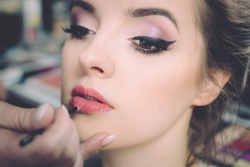 A close-up of a woman having makeup applied to her lips. Her eyes are accentuated with dark eyeliner and pink eye shadow. The focus is on her face, especially the eyes and lips, while the background is blurred, suggesting a makeup studio environment.