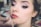 A close-up of a woman having makeup applied to her lips. Her eyes are accentuated with dark eyeliner and pink eye shadow. The focus is on her face, especially the eyes and lips, while the background is blurred, suggesting a makeup studio environment.