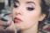 A close-up of a woman having makeup applied to her lips. Her eyes are accentuated with dark eyeliner and pink eye shadow. The focus is on her face, especially the eyes and lips, while the background is blurred, suggesting a makeup studio environment.