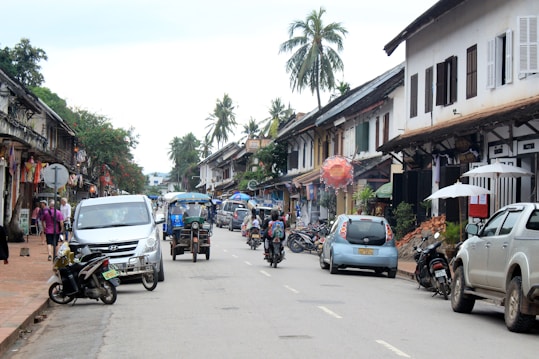 A street scene features a variety of vehicles including cars, motorcycles, and a tuk-tuk moving along a road lined with traditional Southeast Asian architecture. The buildings display a mix of colonial and local influences, with wooden shutters and decorative elements. Palm trees and greenery add a tropical feel, while pedestrians stroll along the sidewalk.