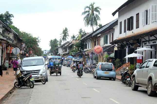 A street scene features a variety of vehicles including cars, motorcycles, and a tuk-tuk moving along a road lined with traditional Southeast Asian architecture. The buildings display a mix of colonial and local influences, with wooden shutters and decorative elements. Palm trees and greenery add a tropical feel, while pedestrians stroll along the sidewalk.