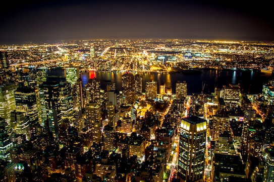 A vibrant cityscape at night with countless illuminated buildings and streets, showcasing a bustling urban environment. The lights from the skyscrapers and city streets create a mesmerizing pattern across the scene, with reflections visible on the body of water in the distance.