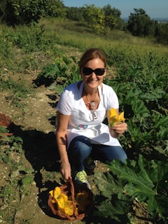 A person is kneeling in a garden or farm field, holding a yellow flower in one hand and a wicker basket with more yellow flowers in the other. Surrounded by green plants and foliage, the person is wearing a white shirt, an apron, jeans, and sunglasses, and is smiling.