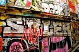 An urban wall covered in vibrant graffiti art, featuring various colors, tags, and designs. The wall is part of a stairwell with metal railings, creating a gritty and artistic atmosphere.