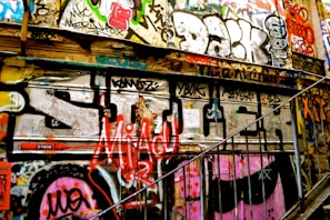 An urban wall covered in vibrant graffiti art, featuring various colors, tags, and designs. The wall is part of a stairwell with metal railings, creating a gritty and artistic atmosphere.