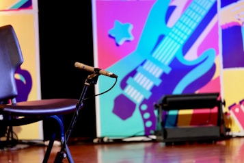 A dynamic, colorful background featuring a stylized guitar in blues and purples with abstract shapes in the background. In the foreground, there's an empty chair and a microphone on a stand, indicating a performance setting.